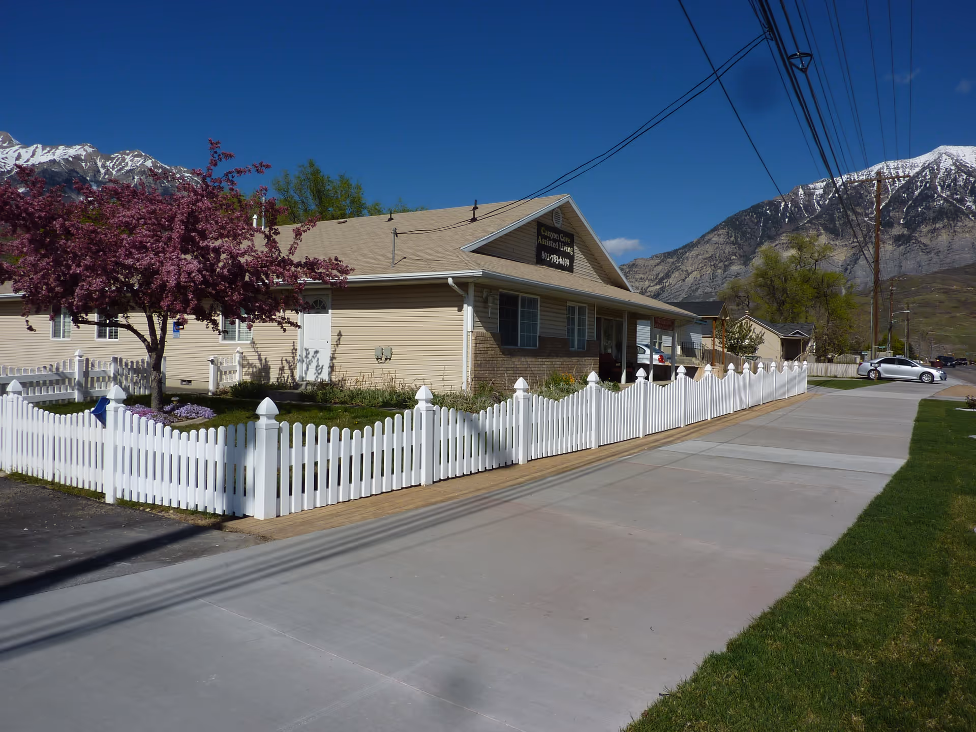 Single-story assisted living building with a white picket fence, a blossoming tree in the yard, and mountains in the background.