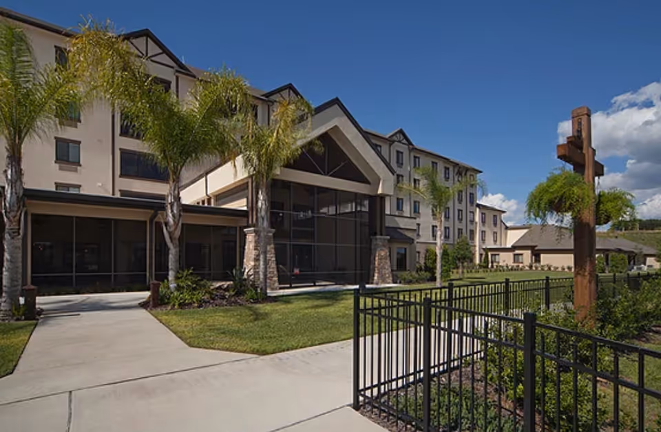 Front entrance of a multi-story senior living building with palm trees, a lawn, and a fenced walkway under a clear blue sky.