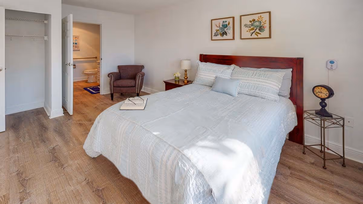 Bright bedroom with a made bed with a wooden headboard, nightstands, an armchair, open closet and visible ensuite bathroom.