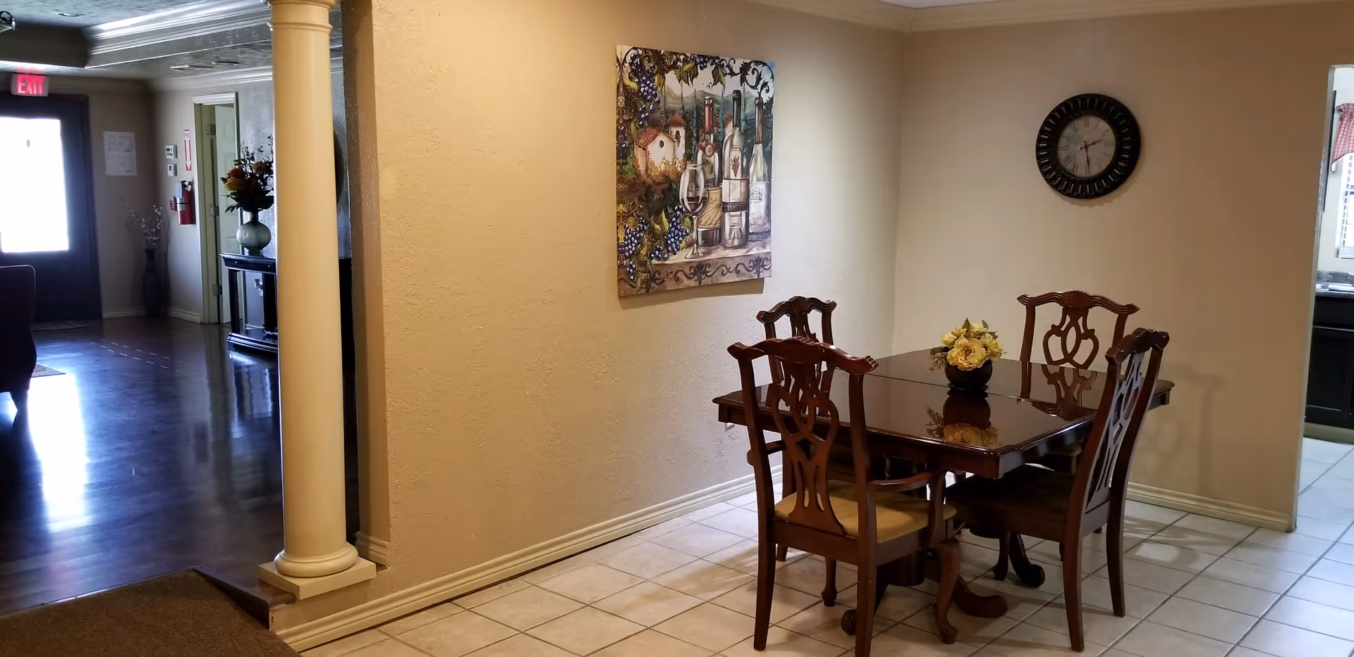 Dining area with a wooden table and four chairs on a tiled floor, a wall clock and painting, and a nearby column leading to a hallway.