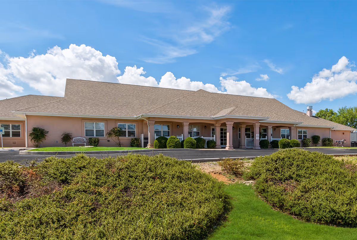 Exterior view of a single-story senior living facility building with a beige facade, multiple windows, a covered entrance with columns, and well-maintained bushes and greenery in the foreground under a blue sky with scattered clouds.