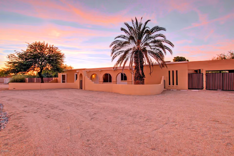 Single-story stucco building with arched windows and a large palm tree in a gravel front yard at sunset.