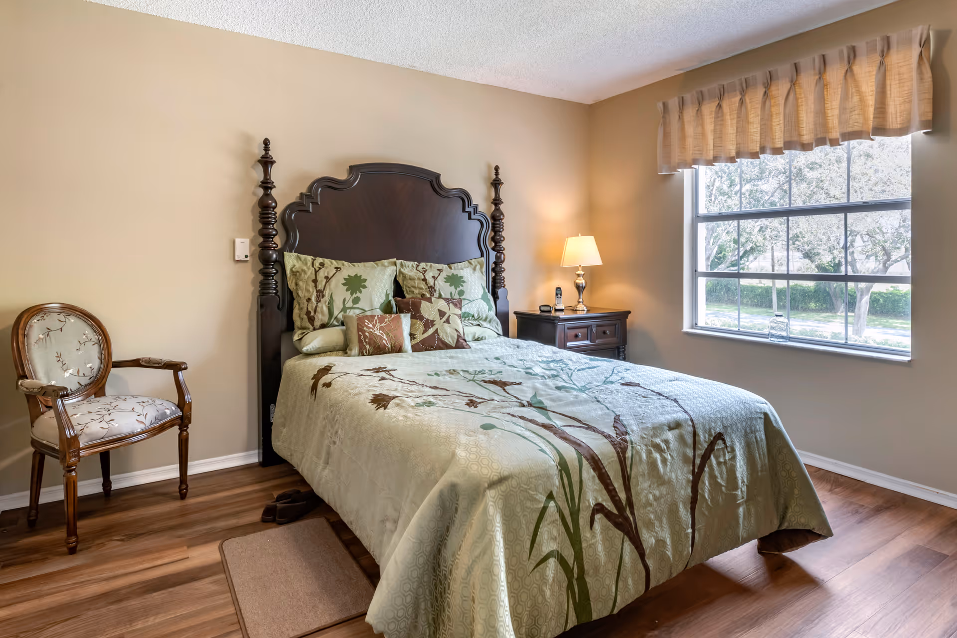A cozy bedroom with a decorative wooden headboard, patterned green bedspread, nightstand and lamp beside a large window.