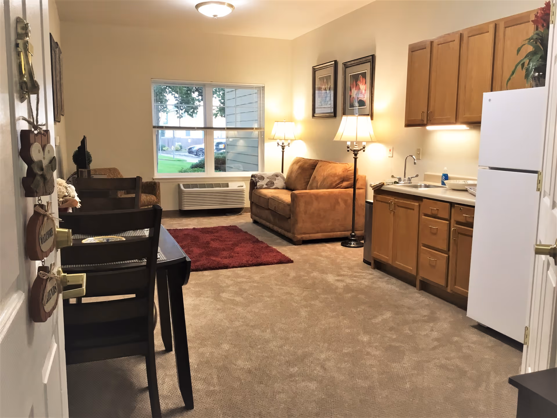 Interior view of a senior living facility apartment featuring a small kitchen area with wooden cabinets, a white refrigerator, and a sink. Adjacent to the kitchen is a living area with a brown couch, two floor lamps, framed floral artwork on the wall, a red rug, and a window showing an outdoor view with greenery and parked cars. A dining table with chairs is partially visible near the entrance door.
