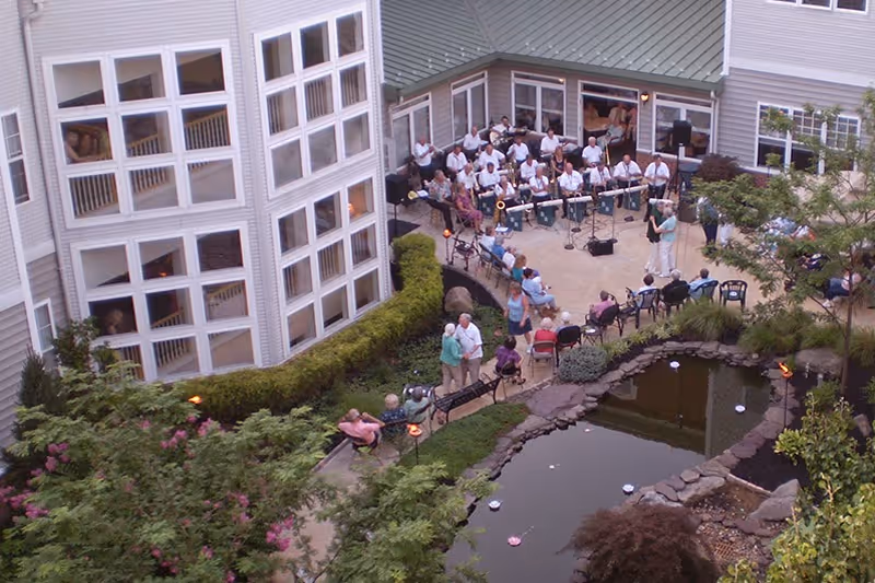 Outdoor courtyard area of a senior living facility with a pond surrounded by rocks and greenery. Several seniors are seated on chairs and benches around the courtyard, watching a band of musicians performing near the building. The building has large windows and light-colored siding.