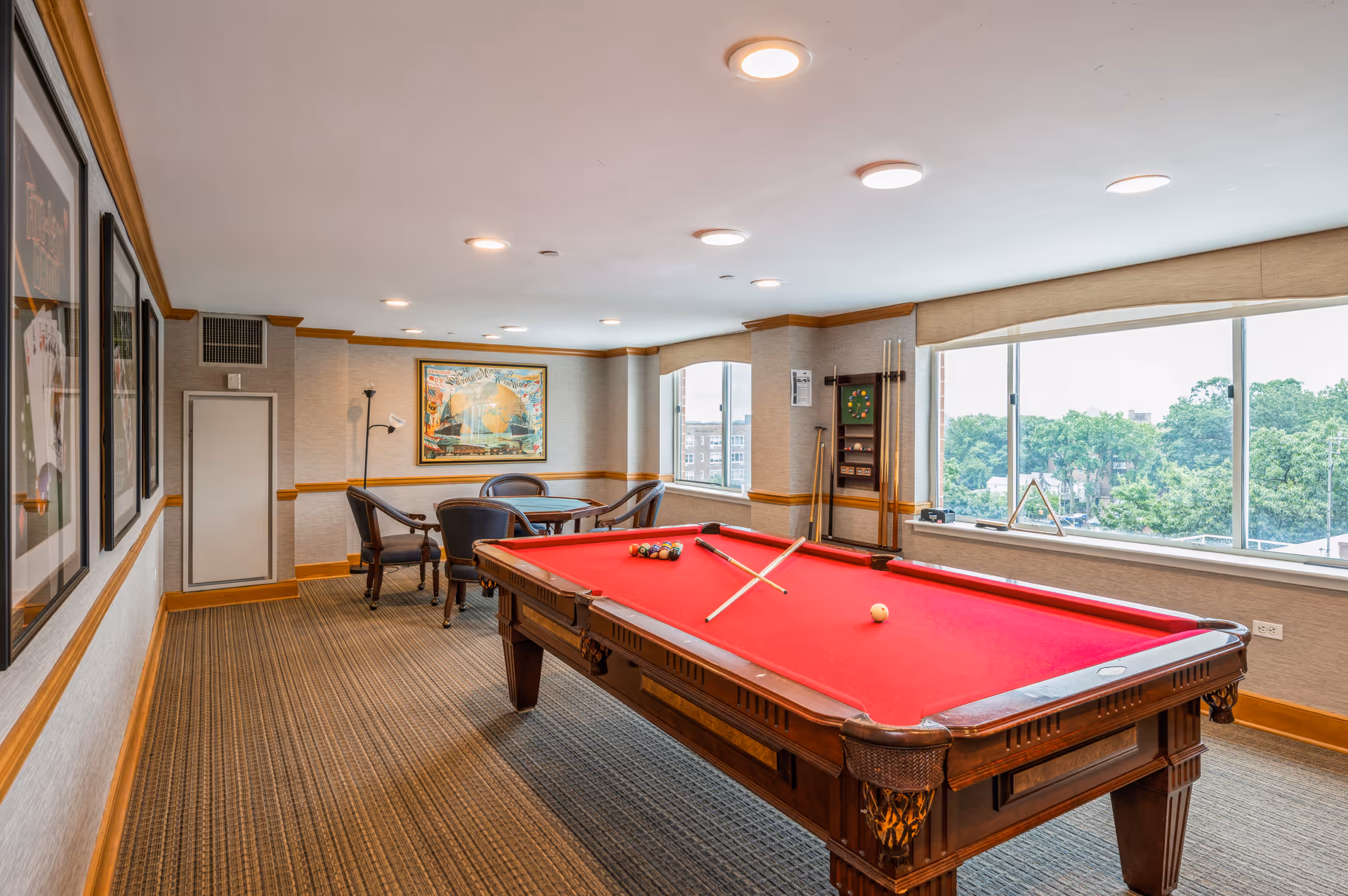 A bright game room with a red felt pool table in the center, two pool cues crossed on the table, and a set of billiard balls arranged. The room has large windows with a view of trees outside, a small round table with four chairs in the corner, framed pictures on the wall, and a rack holding additional pool cues and a dartboard.