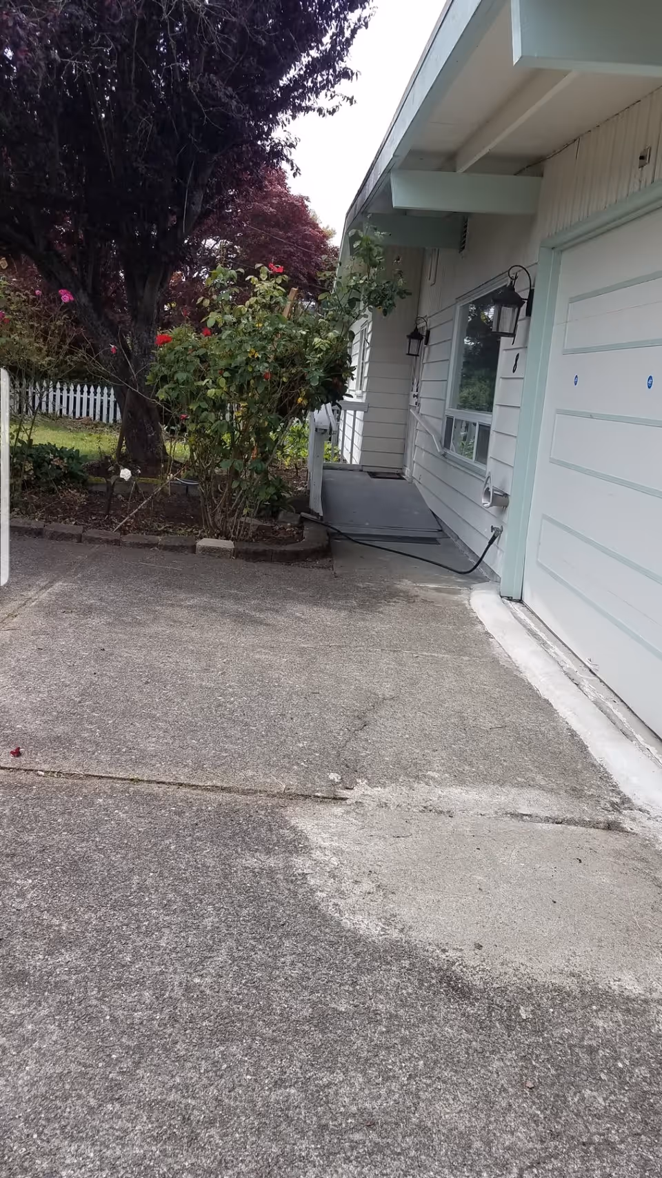 Exterior view of a single-story building with white siding and light green trim. There is a concrete driveway in the foreground, a garden area with bushes and flowers to the left, and a ramp leading to a door on the right side of the building. Two black wall-mounted lanterns are visible near the door and window.