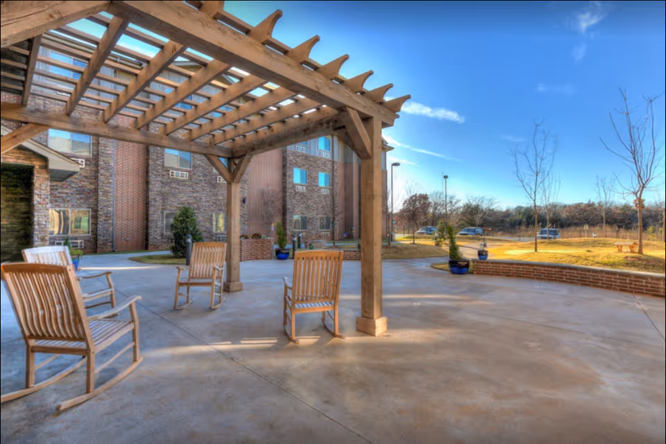 Outdoor patio area at Teal Creek Senior Living with a wooden pergola and several wooden rocking chairs arranged underneath. The background shows a multi-story brick building, some potted plants, a few leafless trees, and a clear blue sky.