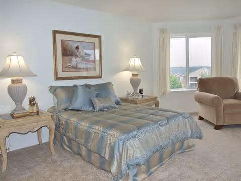 A neatly made bed with a blue and gold patterned comforter and matching pillows in a bedroom. There are two bedside tables with lamps on each side of the bed. A framed picture hangs on the wall above the bed. A beige armchair is positioned near a window with cream-colored curtains, and outside the window, some buildings and greenery are visible.