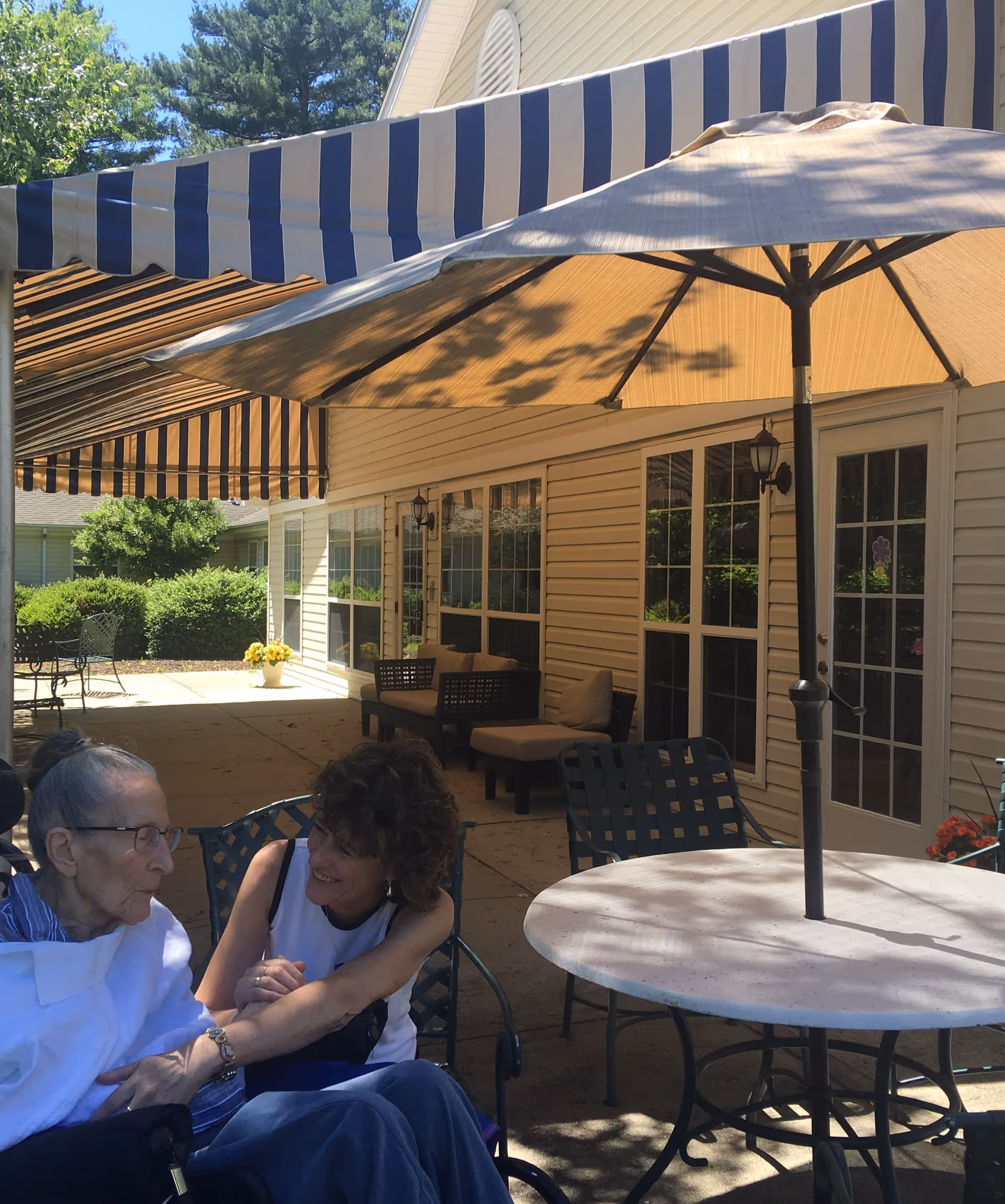 An elderly resident and a caregiver seated under a striped awning and umbrella on a shaded outdoor patio beside the facility building.