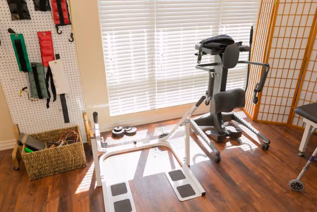 A rehabilitation room with exercise equipment including a standing frame and weights. There is a basket with resistance bands and other therapy tools on the floor, a pegboard with hanging straps on the wall, and a window with blinds letting in natural light. The floor is wooden and there is a folding screen divider in the corner.