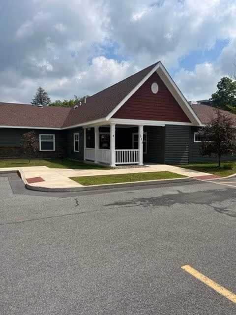 Exterior view of a single-story building with dark green siding and a red gabled roof. The entrance features a small covered porch with white railings. The building is surrounded by a paved parking area and some small trees and grass patches. The sky is partly cloudy.
