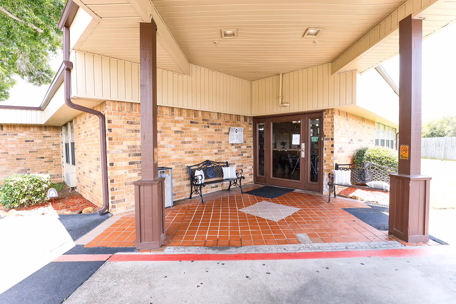 Covered entrance area of a brick building with two brown pillars supporting the roof. There are two black metal benches with cushions on either side of the entrance door. The floor is tiled with reddish-brown tiles and a diamond-shaped pattern in the center. A small garden with bushes is visible on the left side.