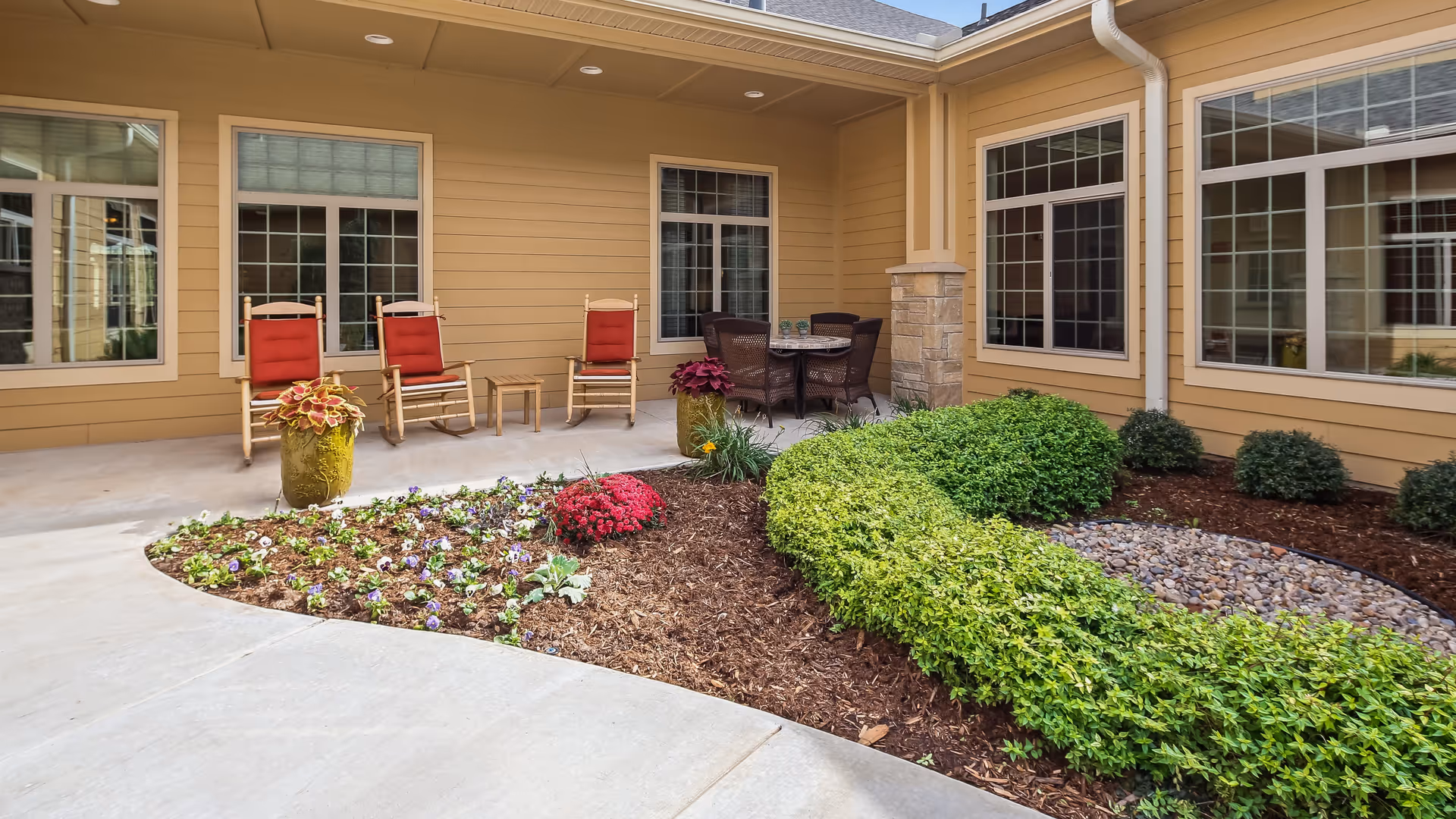 Outdoor patio area at Legend Memory Care at Rivendell featuring a covered seating space with three wooden rocking chairs with red cushions, a small table, and a round table with four wicker chairs. The patio is surrounded by a garden with neatly trimmed bushes, colorful flowers, and decorative plants. The building exterior is beige with multiple large windows.