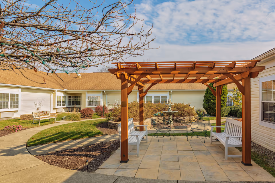Sunny courtyard featuring a wooden pergola with benches, a paved seating area, and a fountain in front of a single-story building.