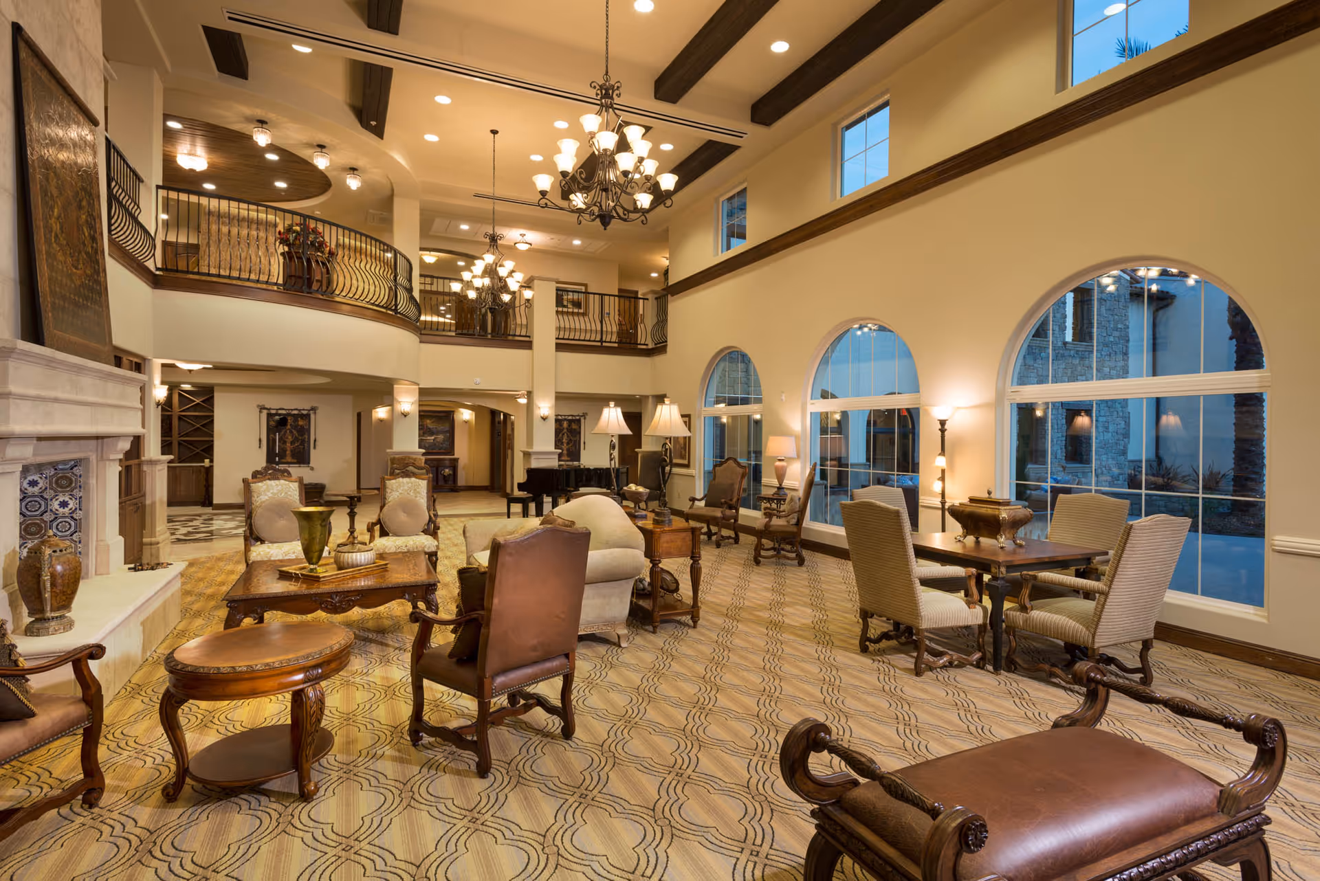 Spacious and elegant senior living facility common area with high ceilings, large arched windows, chandeliers, and classic wooden furniture including armchairs, tables, and a fireplace. The room has a warm and inviting atmosphere with a patterned carpet and a balcony overlooking the space.