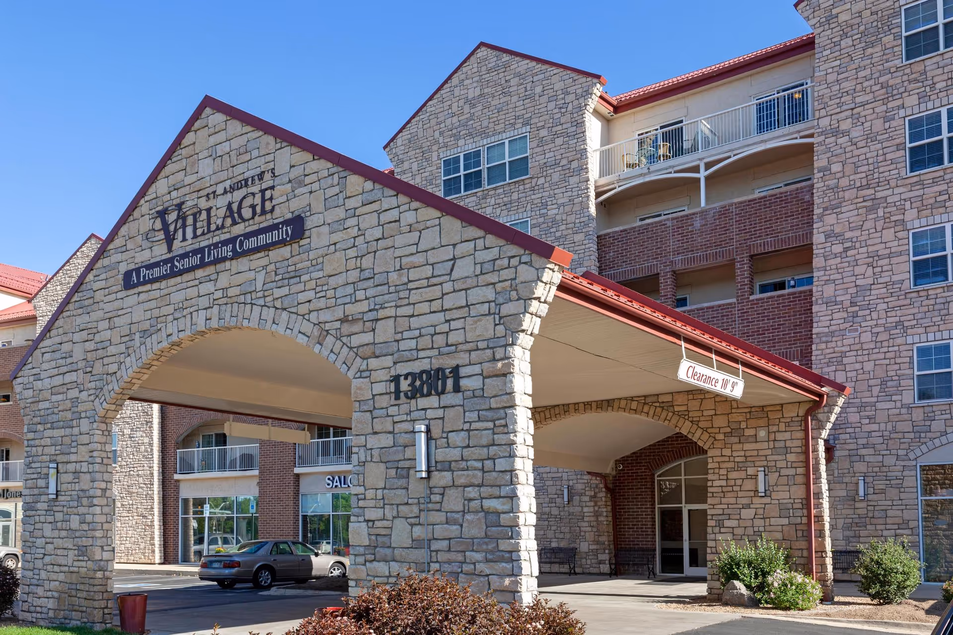 Exterior view of St. Andrew's Village, a senior living community, showing a stone and brick building with a covered entrance and balconies. The entrance has a sign with the community name and the address number 13801. There are some bushes and a car parked near the entrance.