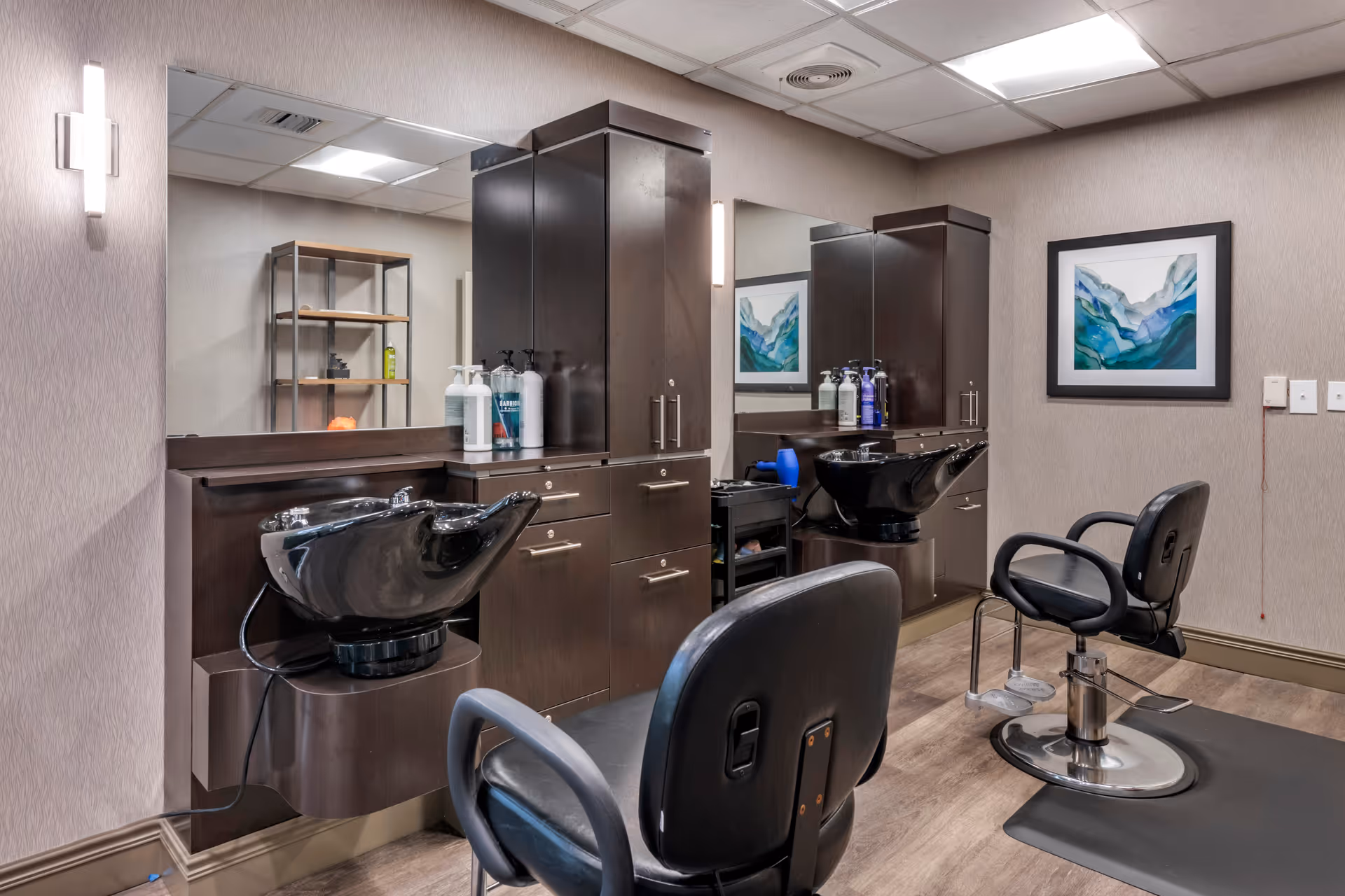 Interior of a hair salon area with two black salon chairs and two black hair washing sinks mounted on dark wood cabinetry. There are large mirrors above the sinks, shelves with hair products, and a framed abstract painting on the wall. The room has beige walls and a wood floor.