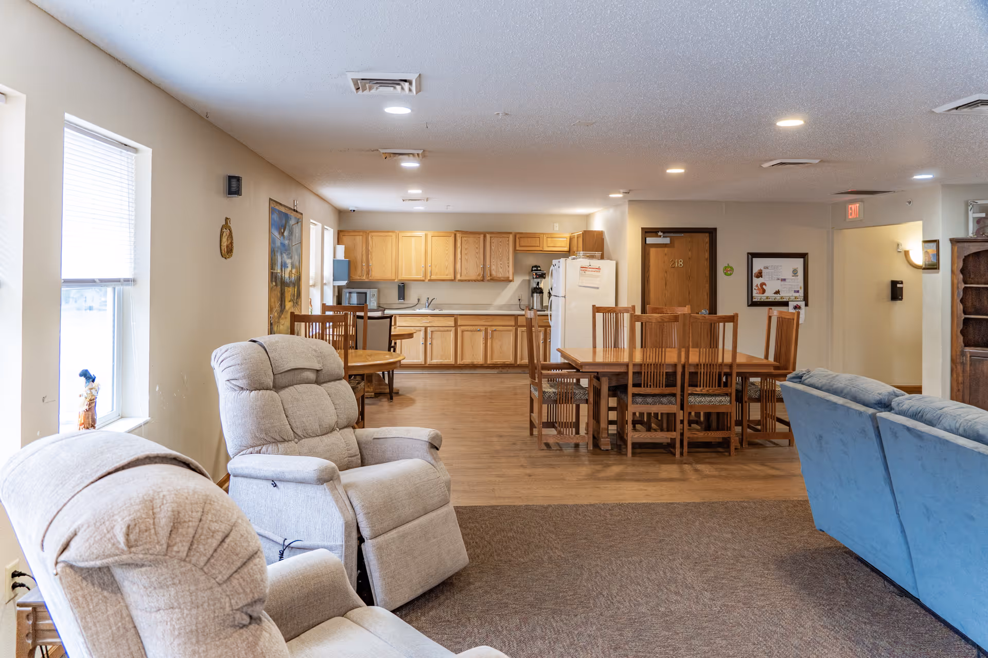 A bright and spacious common area in a senior living facility featuring two beige recliners and a blue sofa in the foreground, a wooden dining table with six chairs in the middle, and a kitchen area with wooden cabinets, a refrigerator, and a microwave in the background. The room has large windows on the left side letting in natural light, light-colored walls, and ceiling lights.