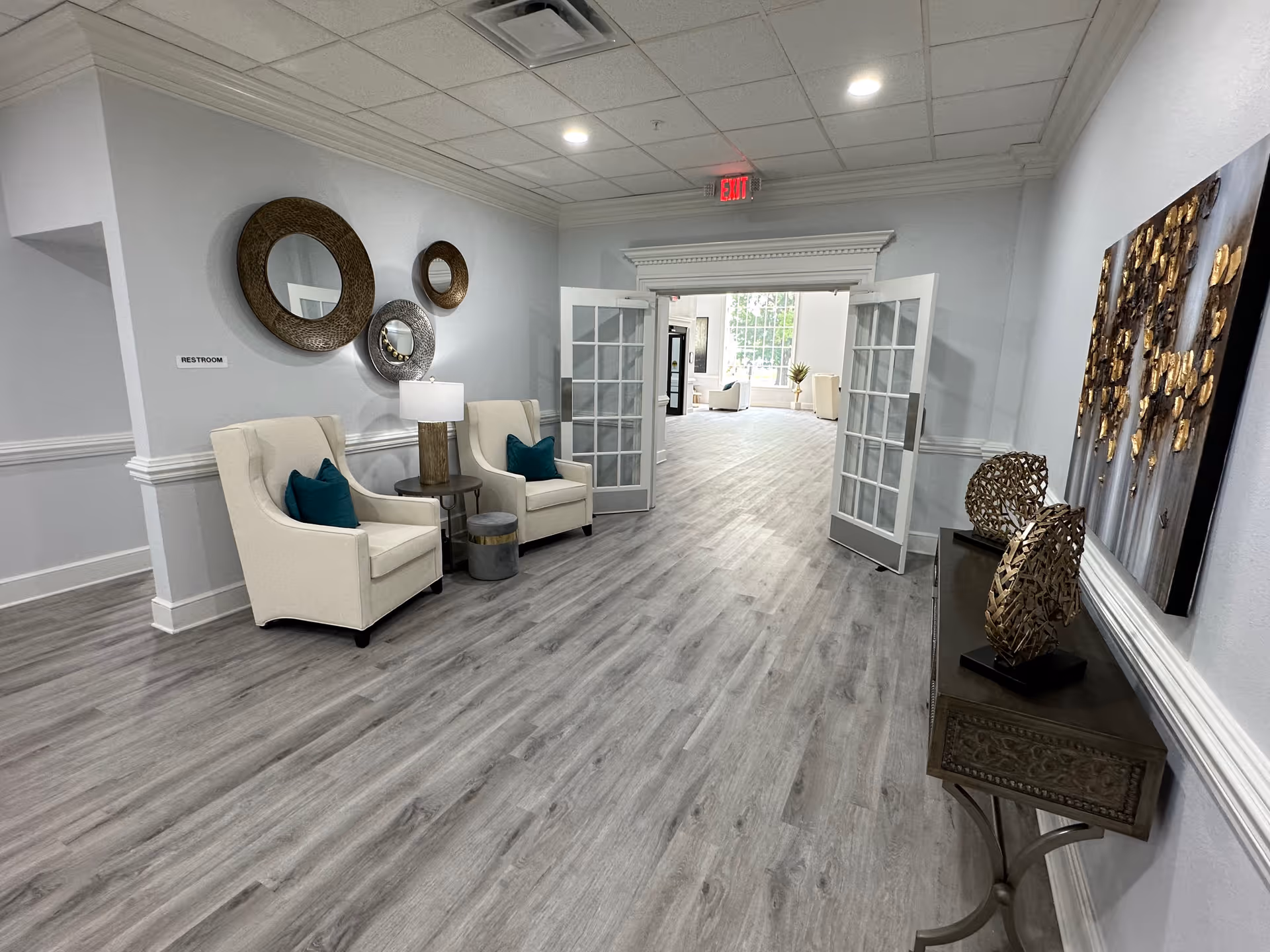 Bright, modern interior hallway and seating area with two armchairs, round wall mirrors, a decorative console table, and open French doors leading to a sunlit lounge.