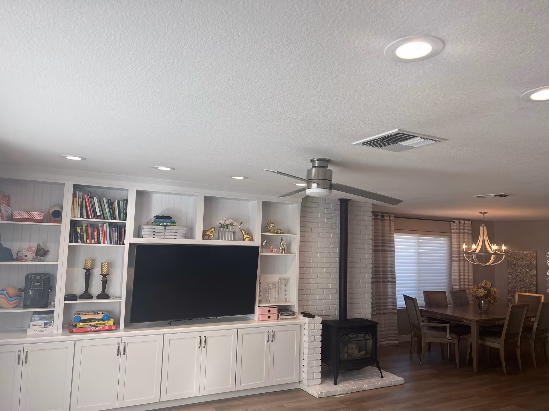 A cozy living and dining area featuring a white built-in entertainment center with shelves holding books, decorative items, and a flat-screen TV. To the right, there is a black wood stove with a white brick surround and a ceiling fan above. In the background, a dining table with chairs is illuminated by a chandelier, with a window covered by striped curtains.