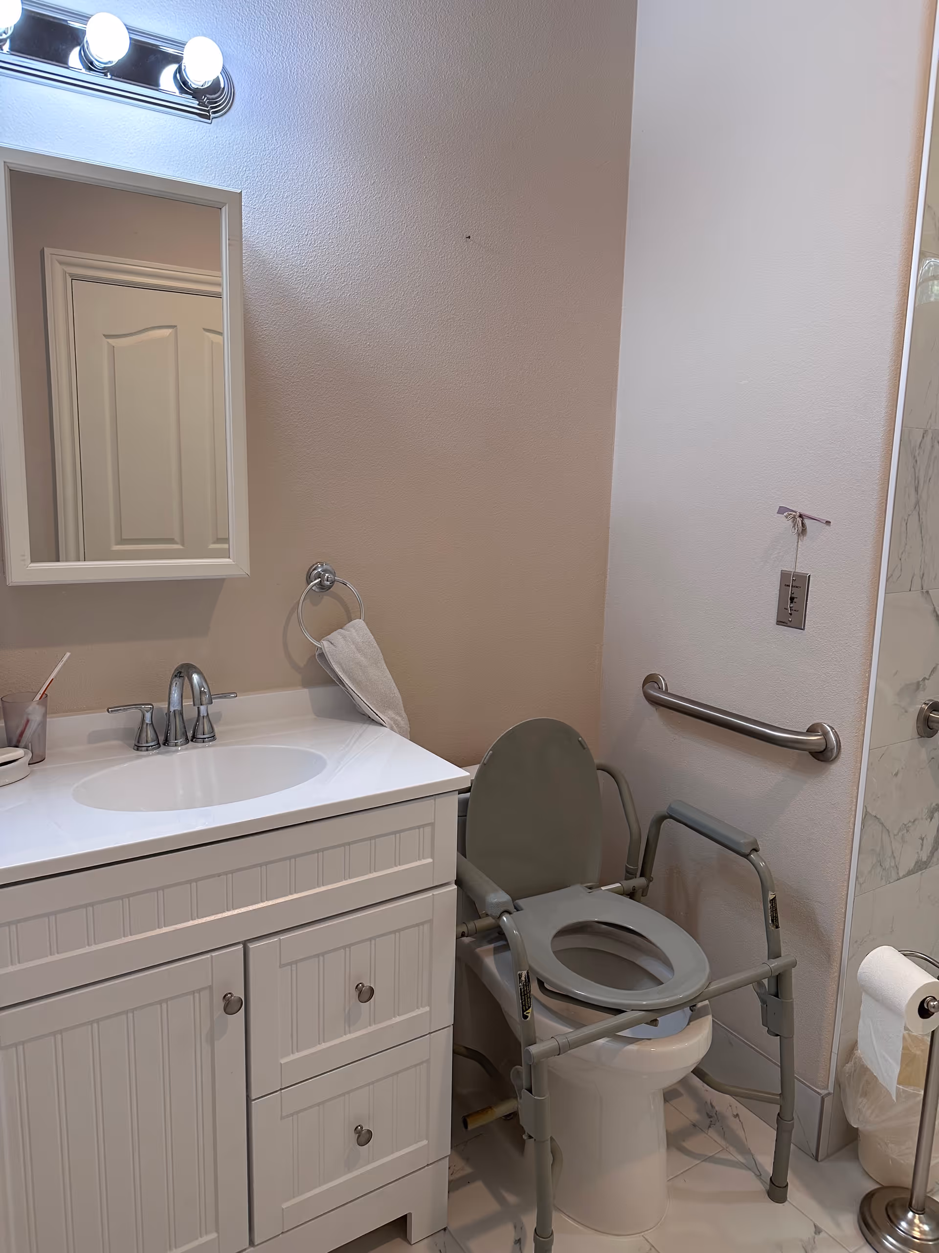 Bathroom with a white vanity and mirror, a raised toilet seat with a support frame and grab bar, and part of a shower visible.