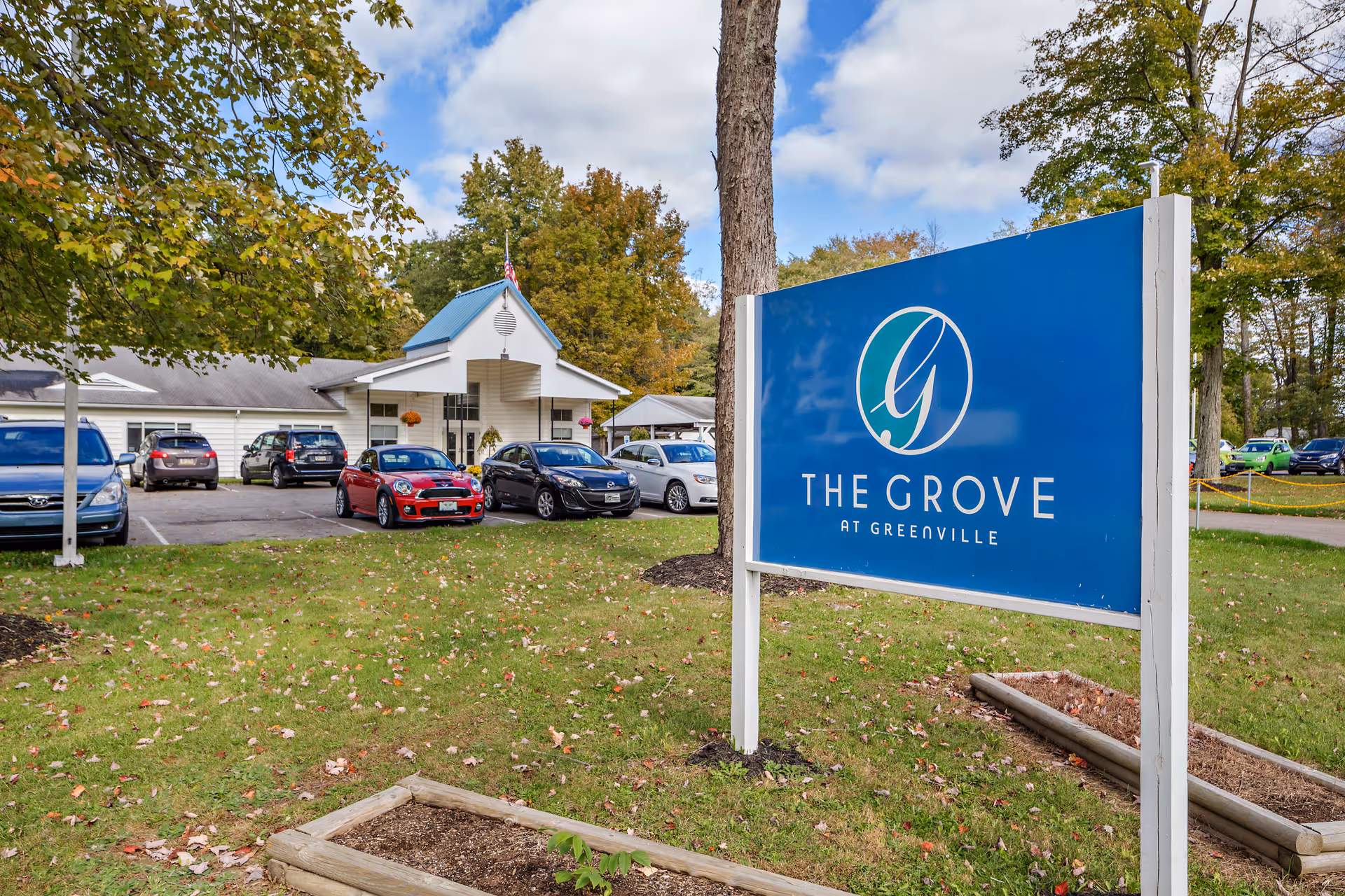 Exterior view of The Grove at Greenville facility with a blue sign displaying the facility name in the foreground. Several cars are parked in front of a single-story building surrounded by trees with autumn foliage.