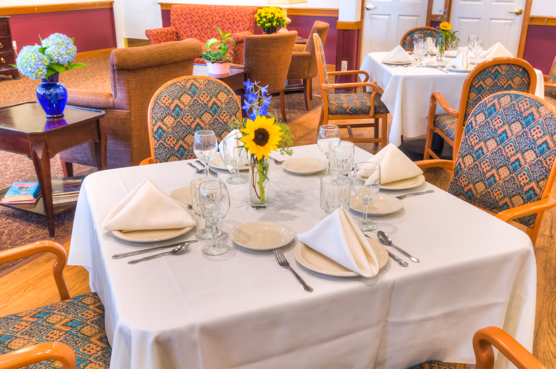 A dining room table set for four with white tablecloth, folded white napkins, plates, glasses, and silverware. A small vase with a sunflower and blue flowers is in the center of the table. In the background, there are upholstered chairs, a side table with a blue vase holding flowers, and a red patterned sofa.