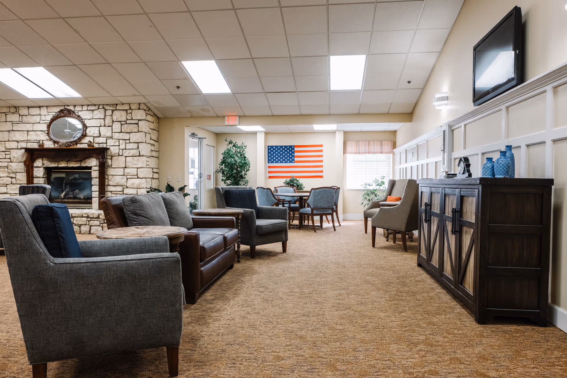A spacious senior living common area with sofas, armchairs, a stone fireplace, and an American flag on the far wall.
