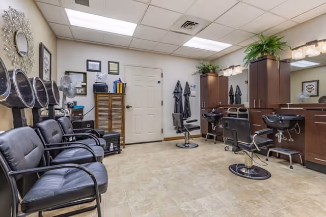 Interior view of a salon area with black salon chairs, hair washing stations, mirrors, and cabinets. The room has a tiled floor, a white door, and a ceiling with fluorescent lights. There are plants on top of the cabinets and some framed pictures on the walls.