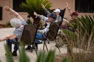 A group of elderly people sitting outdoors on chairs arranged in a row, stretching their arms to the side as part of a group exercise or activity, with plants and a building in the background.