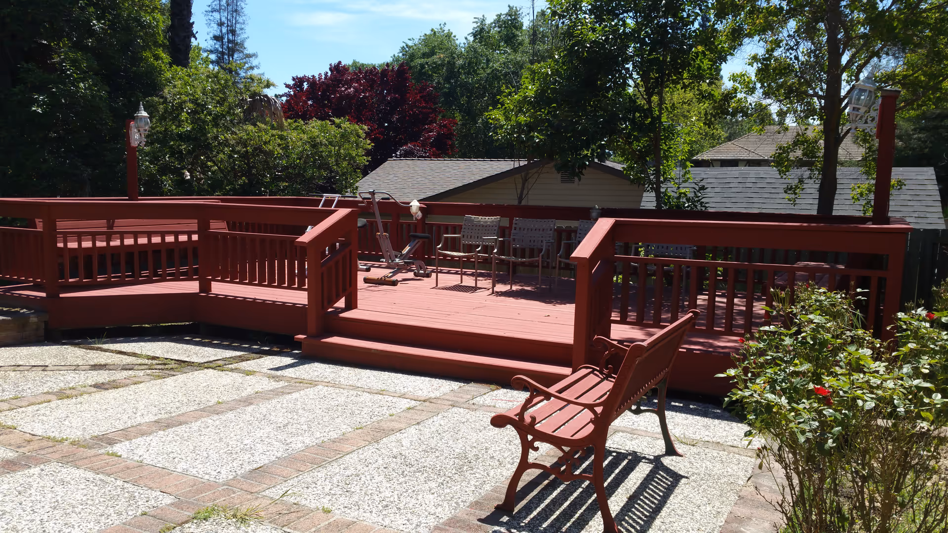 Outdoor patio area with a red wooden deck featuring two chairs and an exercise machine. In front of the deck is a red bench on a paved surface with plants and trees surrounding the area under a clear blue sky.