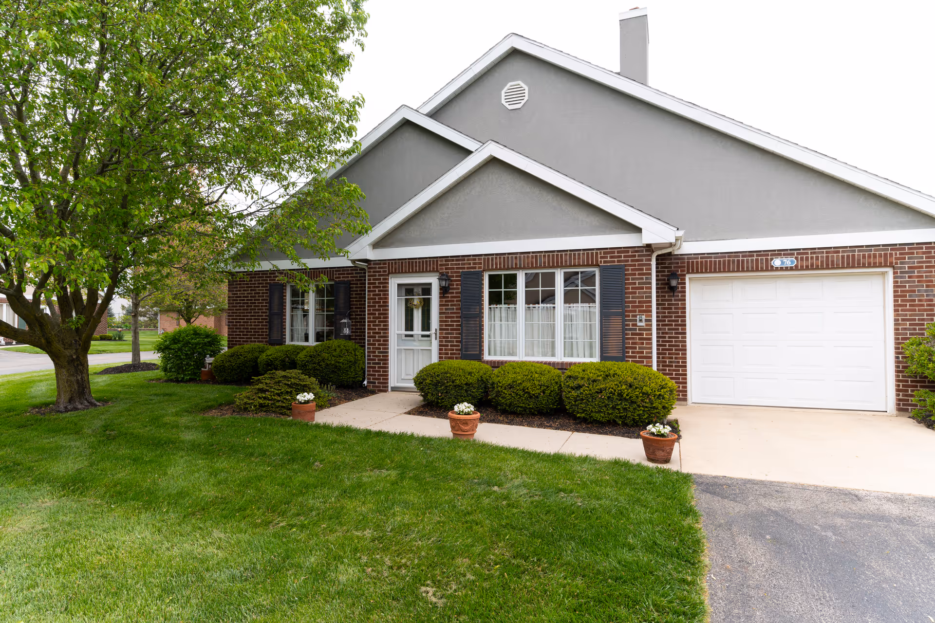 Brick and gray-sided single-story residence with an attached garage, front door, shrubs, potted flowers, driveway and a tree on a green lawn.