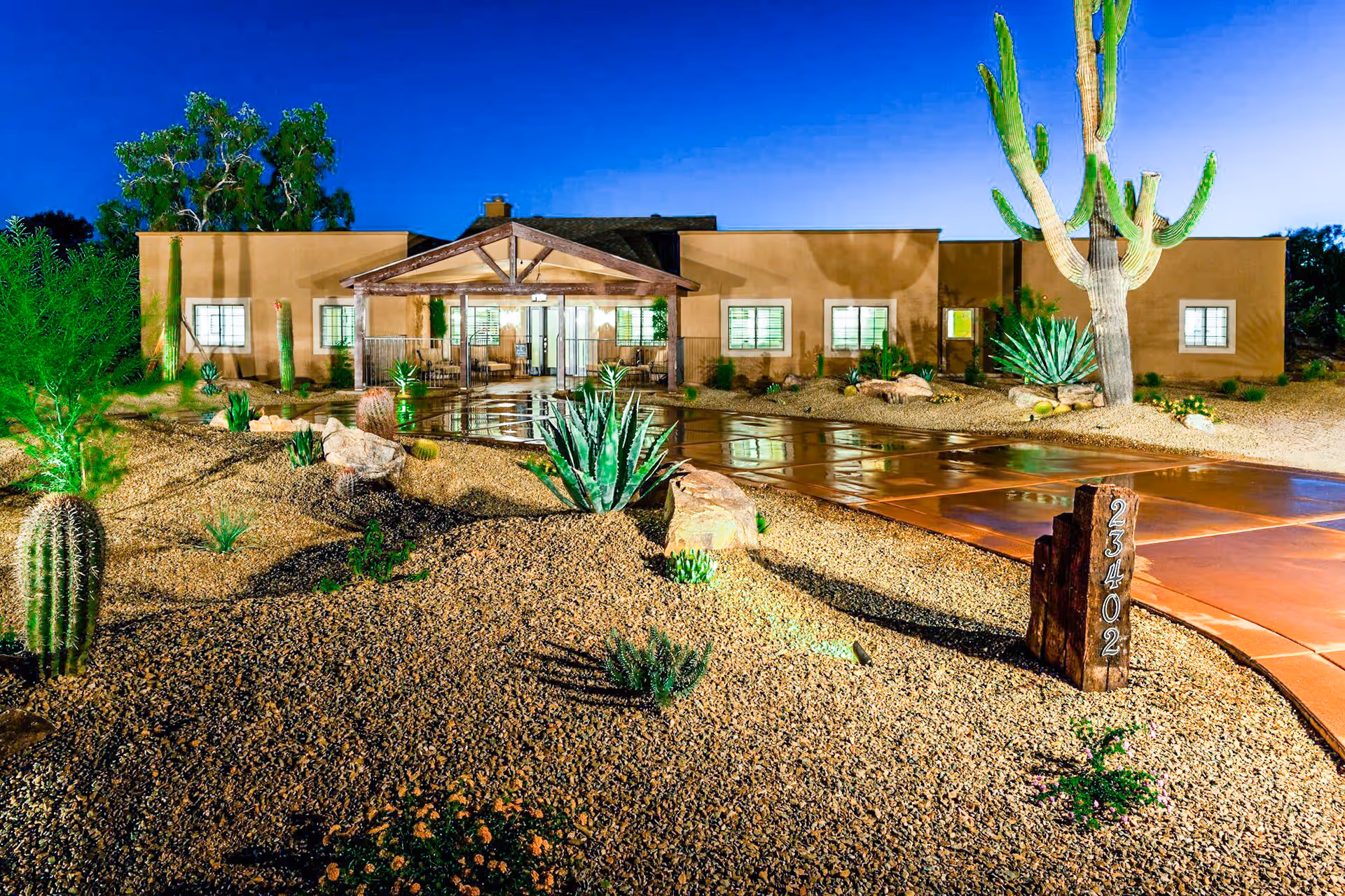 Night view of a single-story desert-style building with a covered entrance and large windows, surrounded by a landscaped yard featuring various cacti, desert plants, and gravel. A wooden post with the number 23402 is visible near a paved walkway leading to the entrance.