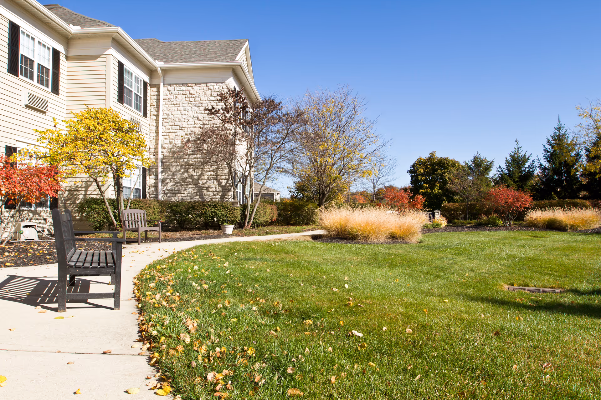 Outdoor area of a senior living facility with a well-maintained lawn, a curved concrete walkway, two wooden benches, and various trees and shrubs with autumn foliage under a clear blue sky.
