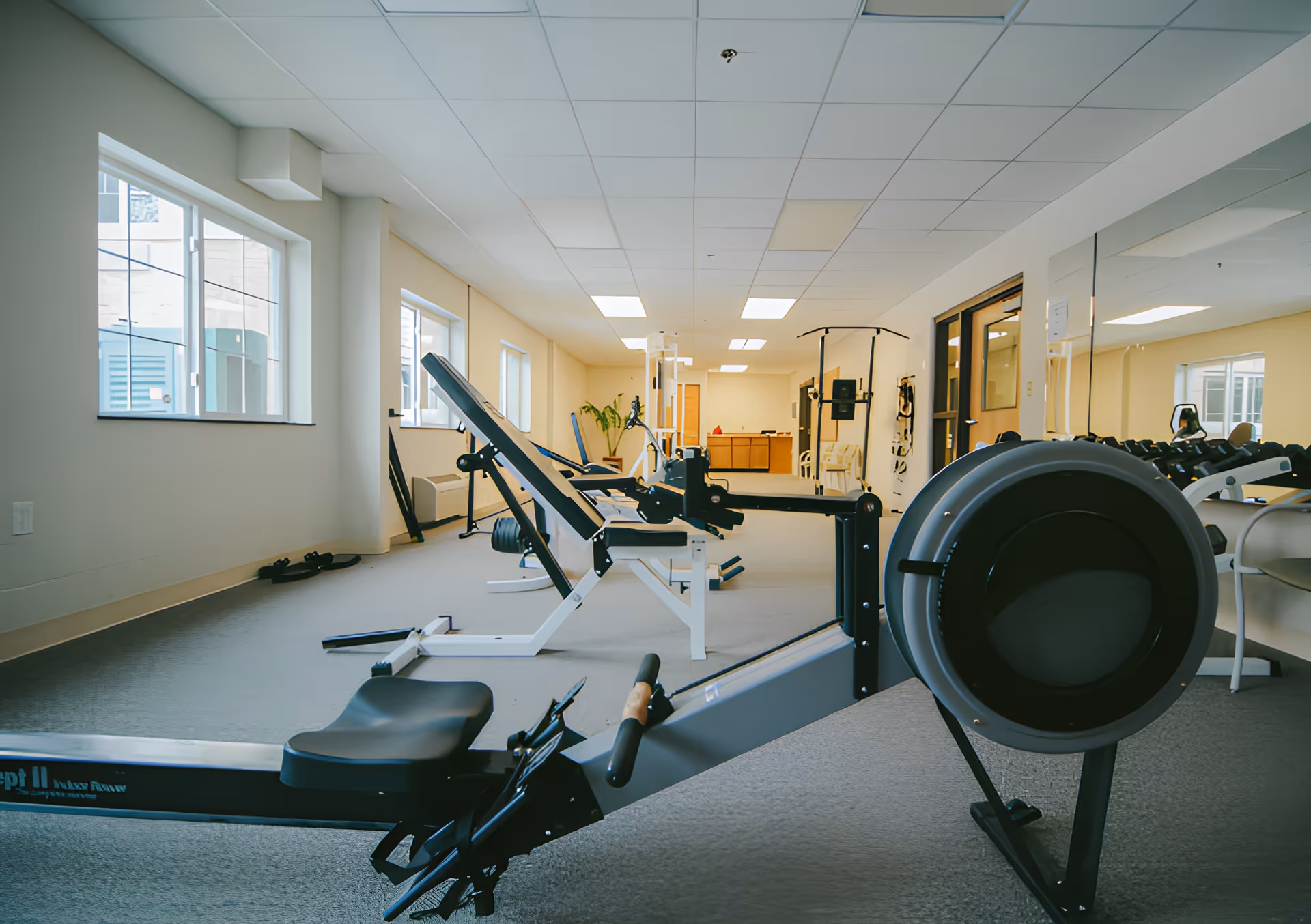 A bright fitness room with exercise equipment including a rowing machine, weight bench, and dumbbells. The room has large windows on the left side letting in natural light, a mirrored wall on the right, and a beige carpeted floor.
