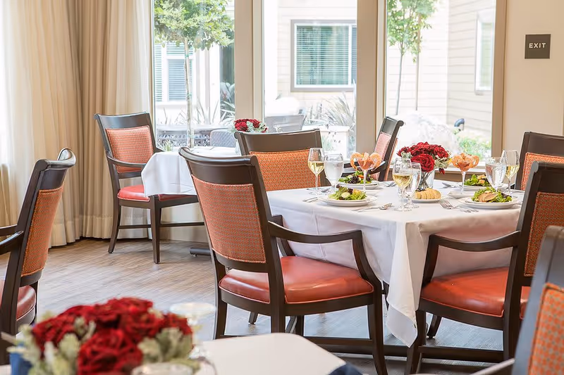 Bright dining room with a table set for a meal, chairs, floral centerpieces and large windows.