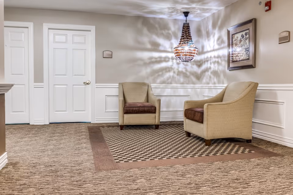 A small seating area in a hallway with two beige armchairs with brown cushions placed on a patterned area rug. The walls are light beige with white wainscoting, and there are two white doors with room number plaques above them. A decorative chandelier hangs from the ceiling, casting patterned shadows on the walls. A framed floral artwork is mounted on the wall to the right.