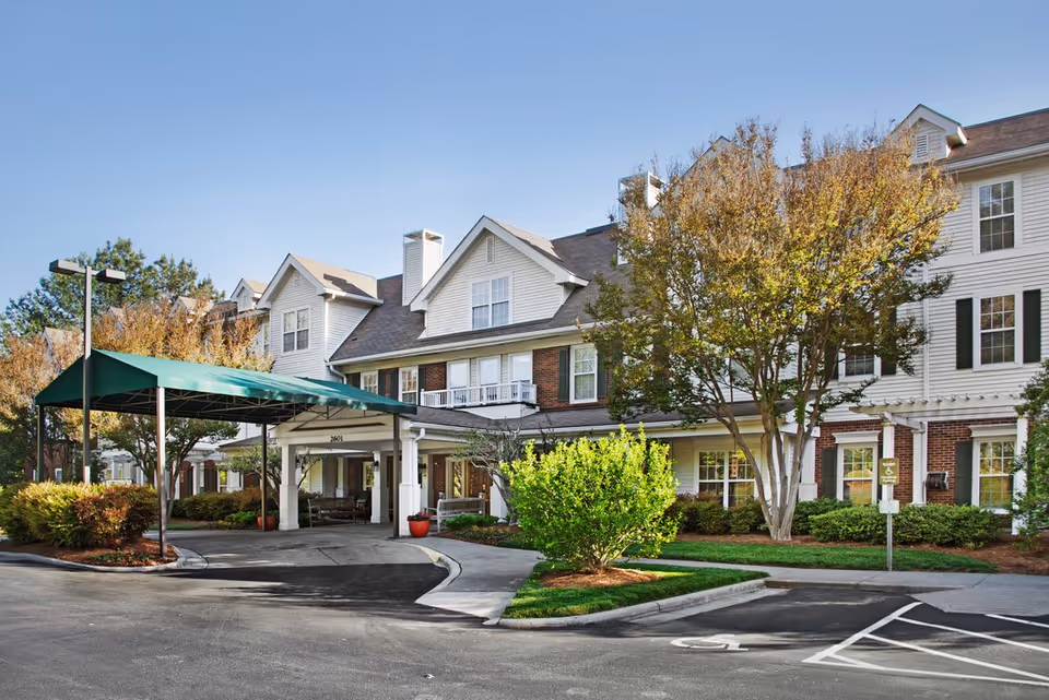 Exterior view of Brighton Gardens of Winston-Salem, showing a multi-story building with white siding and brick accents. The entrance has a covered driveway with a green canopy, surrounded by landscaped bushes and trees under a clear blue sky.