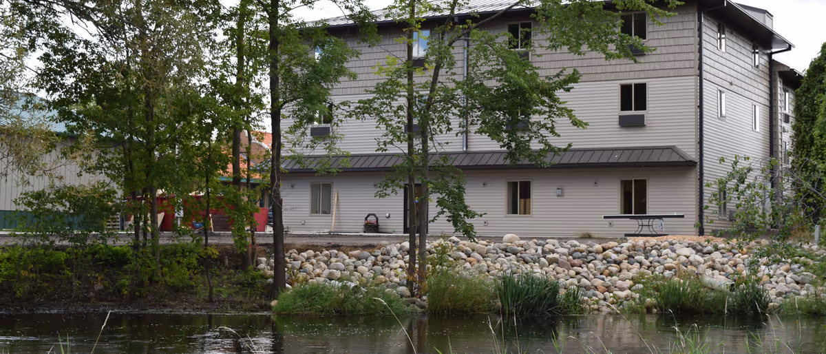 A multi-story building with beige siding and several windows, situated behind a small pond with rocks and greenery along the water's edge. Trees partially obscure the view of the building.