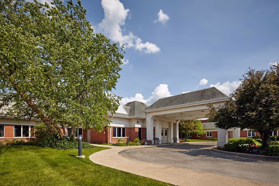 Front entrance of a single-story brick healthcare building with a covered drop-off area, driveway, lawn and trees under a blue sky.