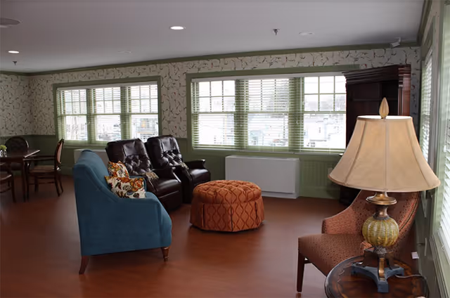 A cozy living room area with large windows featuring green blinds. The room has a blue upholstered sofa with patterned pillows, two dark brown leather recliners, and an orange patterned ottoman in the center. There is also a side table with a decorative lamp and an upholstered chair next to it. The walls have a floral wallpaper with a green wainscoting, and the floor is wooden.