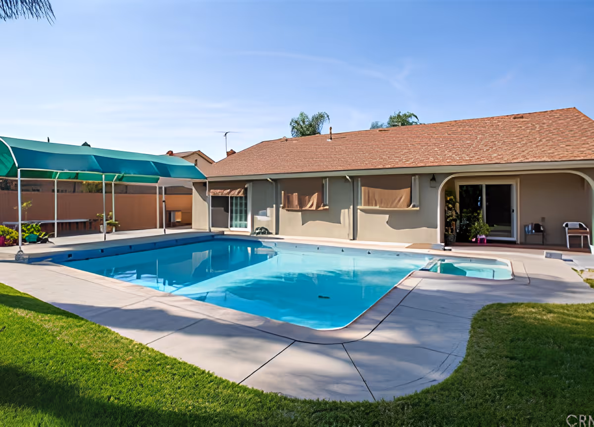 Outdoor swimming pool with clear blue water next to a single-story building with a brown shingled roof. The pool area is surrounded by a concrete deck and green grass. There is a green canopy structure on one side of the pool providing shade. The sky is clear and blue.