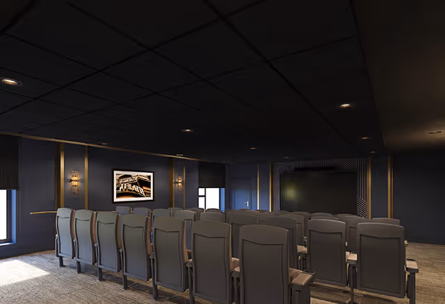 A small theater or presentation room with several rows of gray cushioned chairs facing a large screen on the front wall. The room has dark walls with gold accents, a black ceiling with recessed lighting, and a framed picture on one side wall.