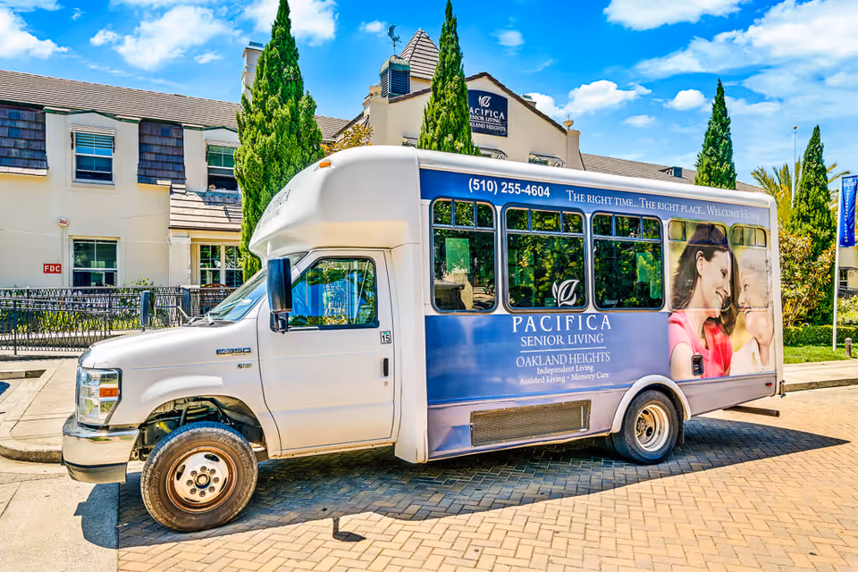 A white and blue shuttle bus parked outside a senior living facility building. The bus has signage for Pacifica Senior Living Oakland Heights, offering independent living, assisted living, and memory care. The building and some tall green trees are visible in the background under a partly cloudy sky.