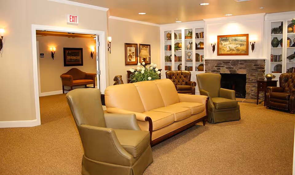 A cozy living room area in Barathaven Alzheimer's Special Care Center featuring a tan leather sofa, two green armchairs, and two brown leather armchairs arranged around a stone fireplace. The walls are decorated with framed artwork and built-in white shelves filled with books and decorative items. Warm lighting fixtures illuminate the space, and an open doorway leads to another room with additional seating.