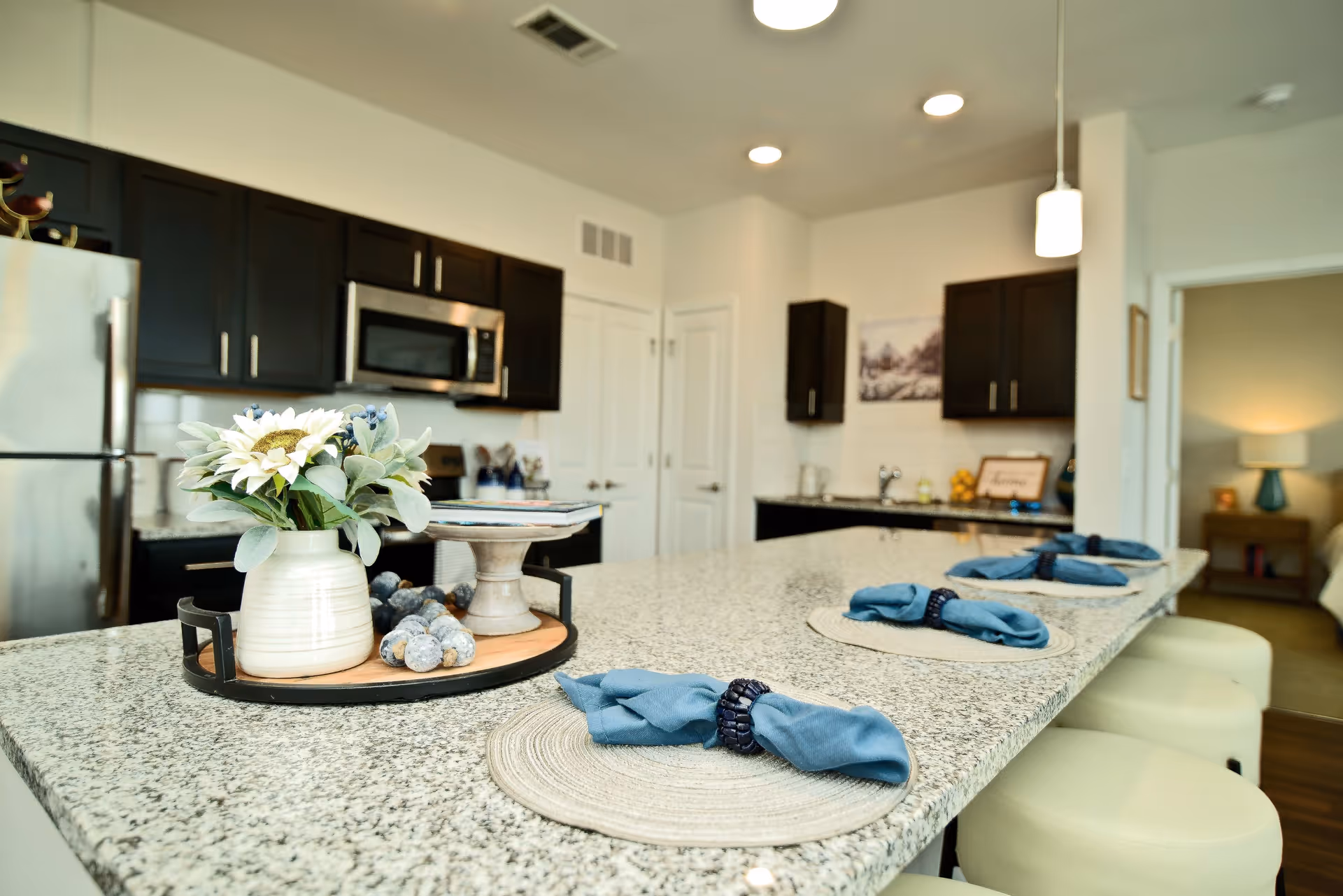 Modern kitchen with a large granite island countertop set with placemats and blue cloth napkins in dark napkin rings. The kitchen features dark wood cabinets, stainless steel refrigerator and microwave, and a decorative tray with a vase of flowers and other items on the island. In the background, a doorway leads to a bedroom with a lit lamp on a nightstand.