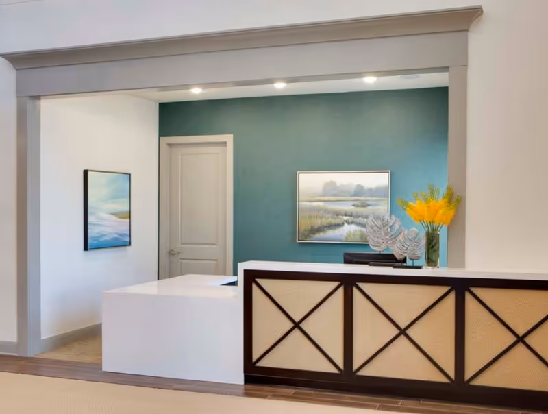 A reception desk in a bright lobby with a teal accent wall, framed artwork, and a vase of yellow flowers.