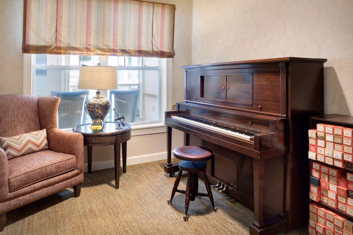 A cozy interior corner featuring a wooden upright piano with a round cushioned piano stool in front. To the left, there is a comfortable upholstered armchair with a chevron-patterned pillow, and a round wooden side table holding a decorative lamp and a small yellow object. A window with a striped valance lets in natural light, and to the right of the piano is a cabinet filled with neatly stacked boxes.
