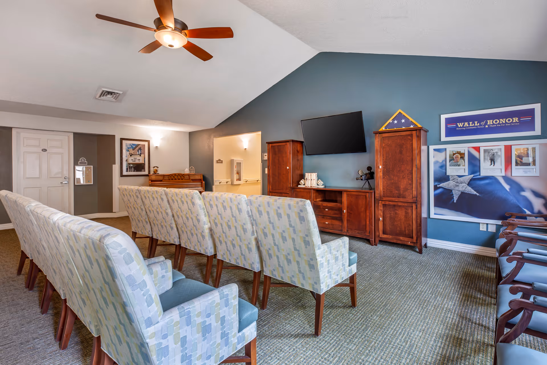 A room with rows of patterned upholstered chairs facing a wooden entertainment center with a flat-screen TV mounted on a blue accent wall. To the right, there is a 'Wall of Honor' display featuring framed photos and an American flag. The room has a ceiling fan, carpeted floor, and a piano against the far wall near a door.